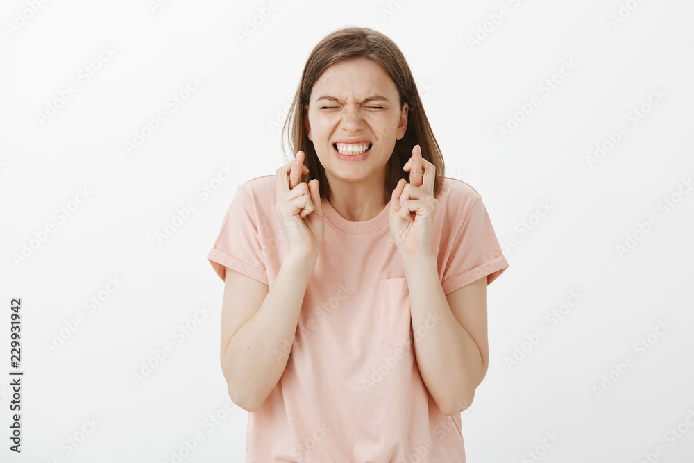 Waist-up shot of troubled cute and funny girl in pink t-shirt, closing eyes and clenching teeth while crossing fingers and begging god help solve problem, making wish or hoping for better Waist-up shot of troubled cute and funny girl in pink t-shirt, closing eyes and clenching teeth while crossing fingers and begging god help solve problem, making wish or hoping for better