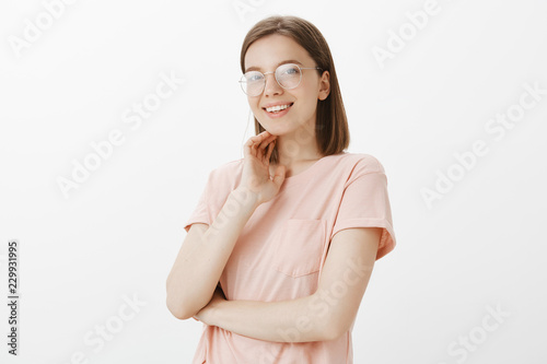 Girl can deal with any task and lead company to success. Portrait of confident carefree and happy woman in glasses, standing half-turned and touching face gently while smiling broadly at camera © Cookie Studio