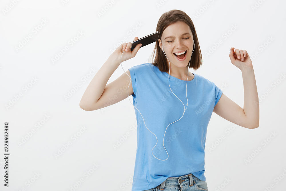 Dancing to the rhythm. Joyful good-looking european woman with tattoos, shaking raised hands and singing along while listening music in earphones and holding smartphone, enjoying great sound Dancing to the rhythm. Joyful good-looking european woman with tattoos, shaking raised hands and singing along while listening music in earphones and holding smartphone, enjoying great sound