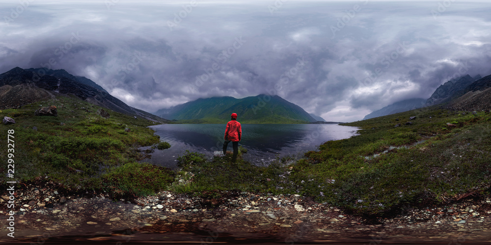 man stands by lake in red jacket in cloudy weather. Spherical panorama 360vr