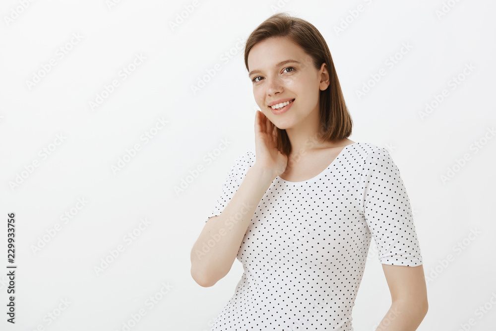 Cute and tender happy european female coworker in casual t-shirt, gently touching neck and smiling joyfully, while standing half-turned over grey wall, being shy and blushing from compliment Cute and tender happy european female coworker in casual t-shirt, gently touching neck and smiling joyfully, while standing half-turned over grey wall, being shy and blushing from compliment