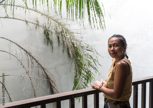 Asian Woman standing on balcony with white wall and tree behind her photo