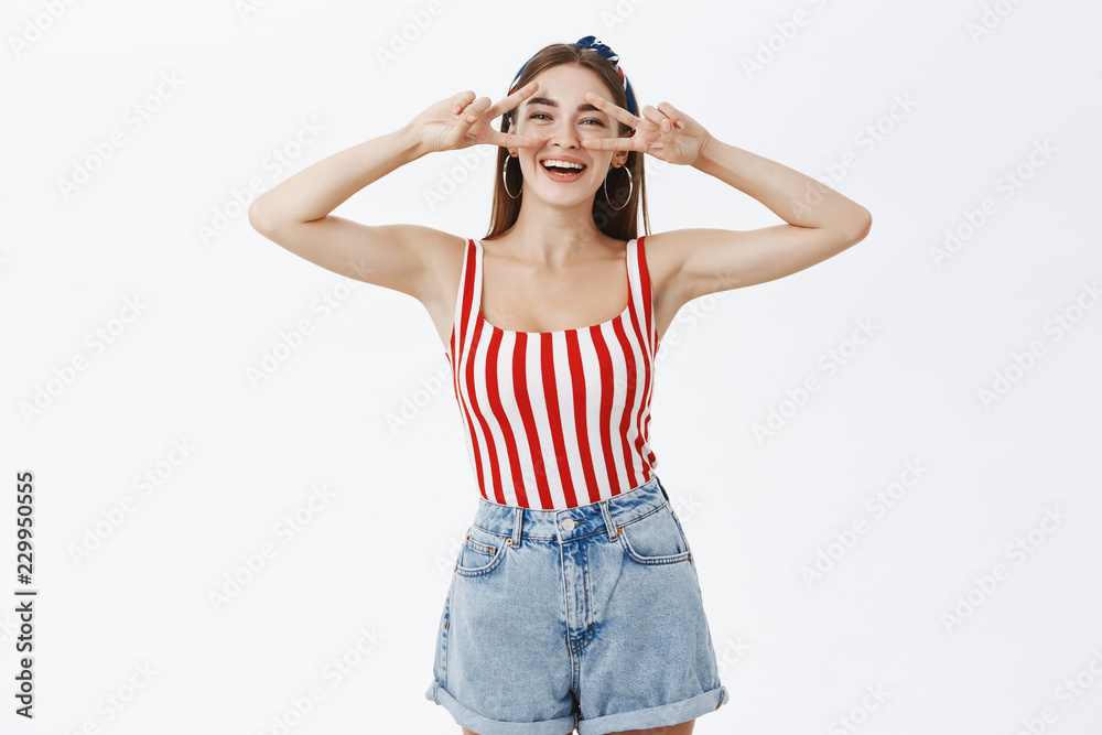Portrait of joyful funny and expressive good-looking woman in pin-up striped top and shorts showing peace signs over eyes and smiling enjoying herself feeling beautiful and inspired over white wall Portrait of joyful funny and expressive good-looking woman in pin-up striped top and shorts showing peace signs over eyes and smiling enjoying herself feeling beautiful and inspired over white wall