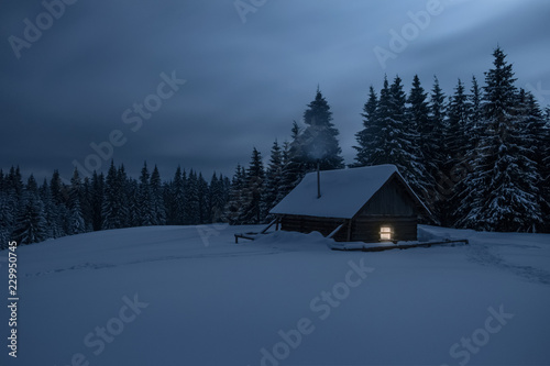 Wooden cabin under stars. Lights shines through the window from inside of the house. Night landscape in winter.