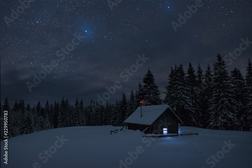Wooden cabin under stars. Lights shines through the window from inside of the house. Night landscape in winter. © ianachyrva