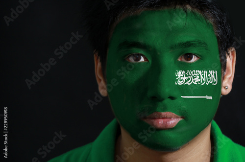 Bilde på lerret Portrait of a man with the flag of the Saudi Arabia painted on his face on black background, a green field with the Shahada or Muslim creed written in the Thuluth script
