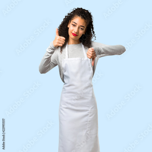 Young girl with apron making good-bad sign. Undecided person between yes or not on isolated blue background