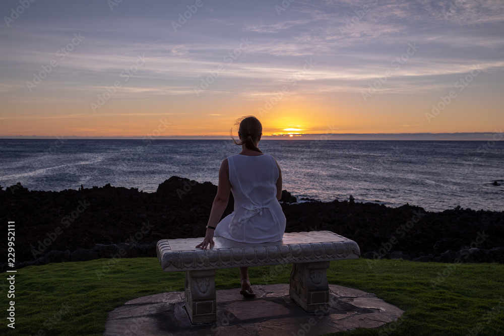 Woman in a white dress sitting on a stone bench watching sunset