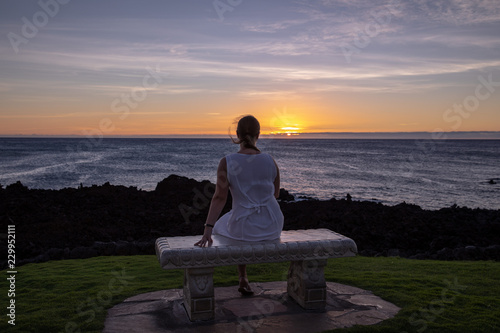 Woman in a white dress sitting on a stone bench watching sunset