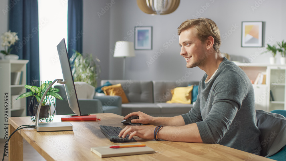Portrait of the Handsome Man Working on Personal Computer while Sitting at His Desk. In the Background Stylish Cozy Living Room. Young Man Playing Computer Games.