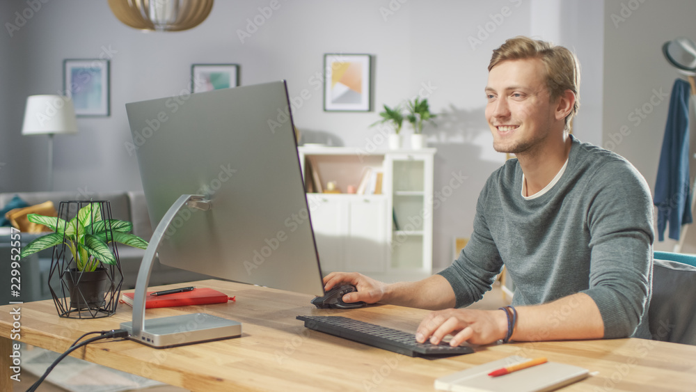 Portrait of the Handsome Man Working on Personal Computer while Sitting at His Desk. In the Background Stylish Cozy Living Room. Young Freelancer Working From Home.