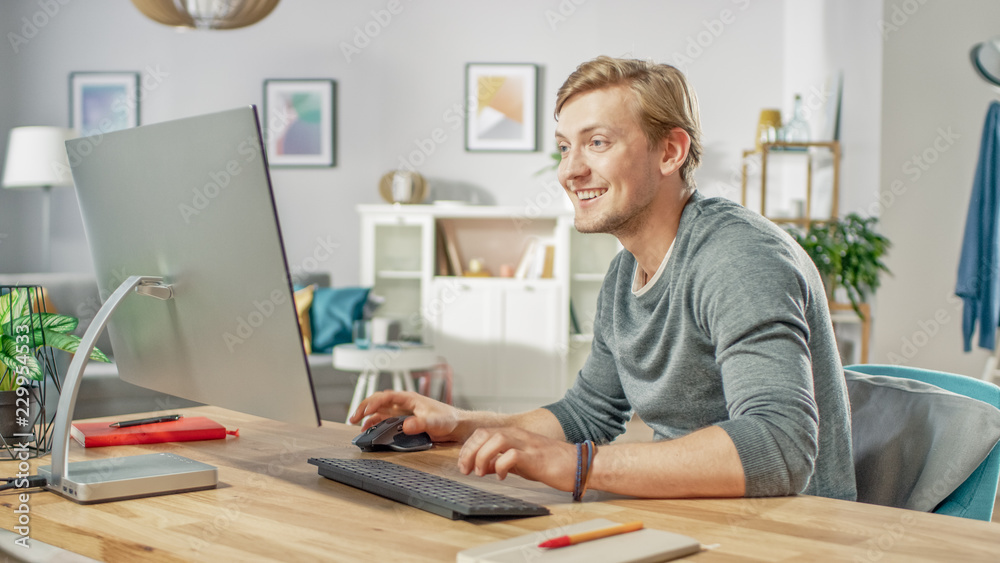 Portrait of the Handsome Smiling Man Working on Personal Computer while Sitting at His Desk. In the Background Stylish Cozy Living Room. Young Freelancer Working From Home. Portrait of the Handsome Smiling Man Working on Personal Computer while Sitting at His Desk. In the Background Stylish Cozy Living Room. Young Freelancer Working From Home.