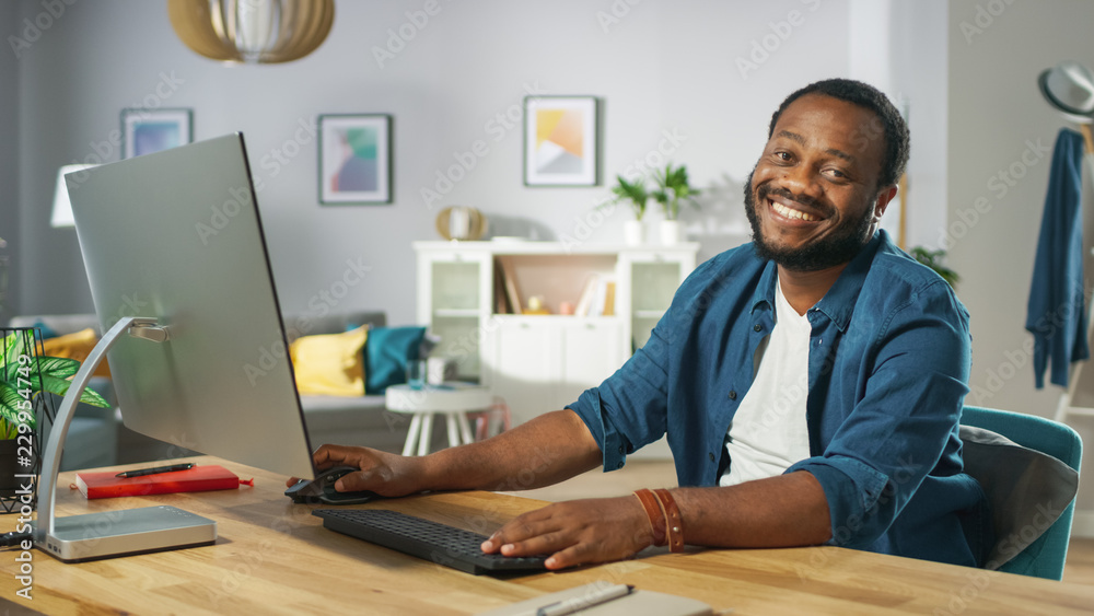 Smiling Young Man Uses Personal Computer, Nods in Agreement and Smiles Charmingly Looking at the Camera. In the Background Cozy Living Room. Smiling Young Man Uses Personal Computer, Nods in Agreement and Smiles Charmingly Looking at the Camera. In the Background Cozy Living Room.