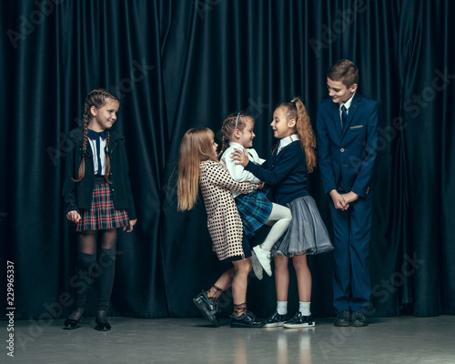 Cute surprised stylish children on dark background. Beautiful stylish teen girls and boy standing together and posing on the school stage in front of the curtain. Classic style. Kids fashion and