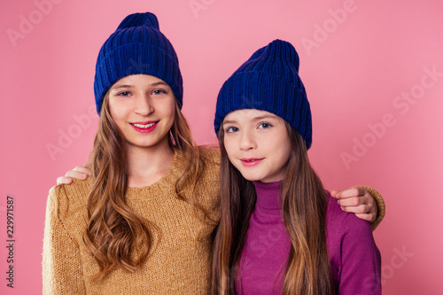 Two beautiful teenage girlfriends hugging and smiling winter wearing knit hats. young female friends sisters portrait in studio pink background.autumn winter fashion look seasonal sale
