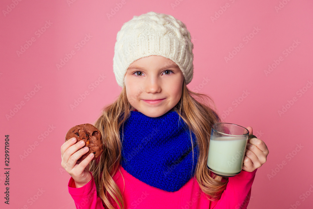 beautiful little girl drinks milk and eats cookies in a knitted hat and scarf on a pink background in the studio,christmas night beautiful little girl drinks milk and eats cookies in a knitted hat and scarf on a pink background in the studio,christmas night