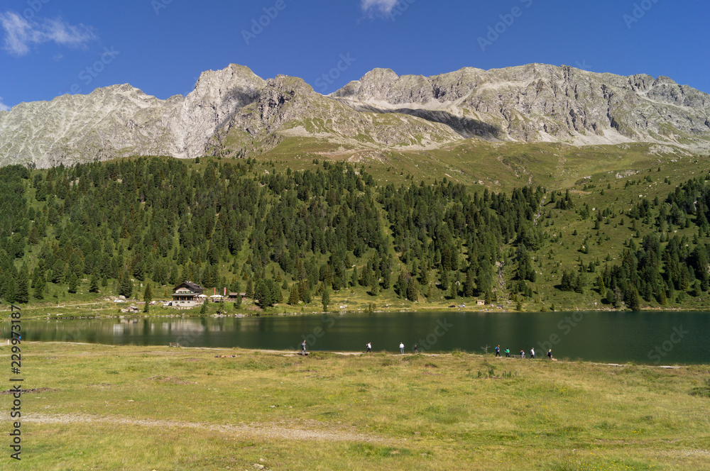 Panorama di un lago di montagna con baita e turisti a passeggio Stock