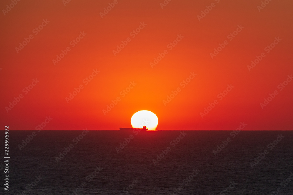 Sunrise from sea water with passing ship on horizon. Red sun disc dawn above the ocean