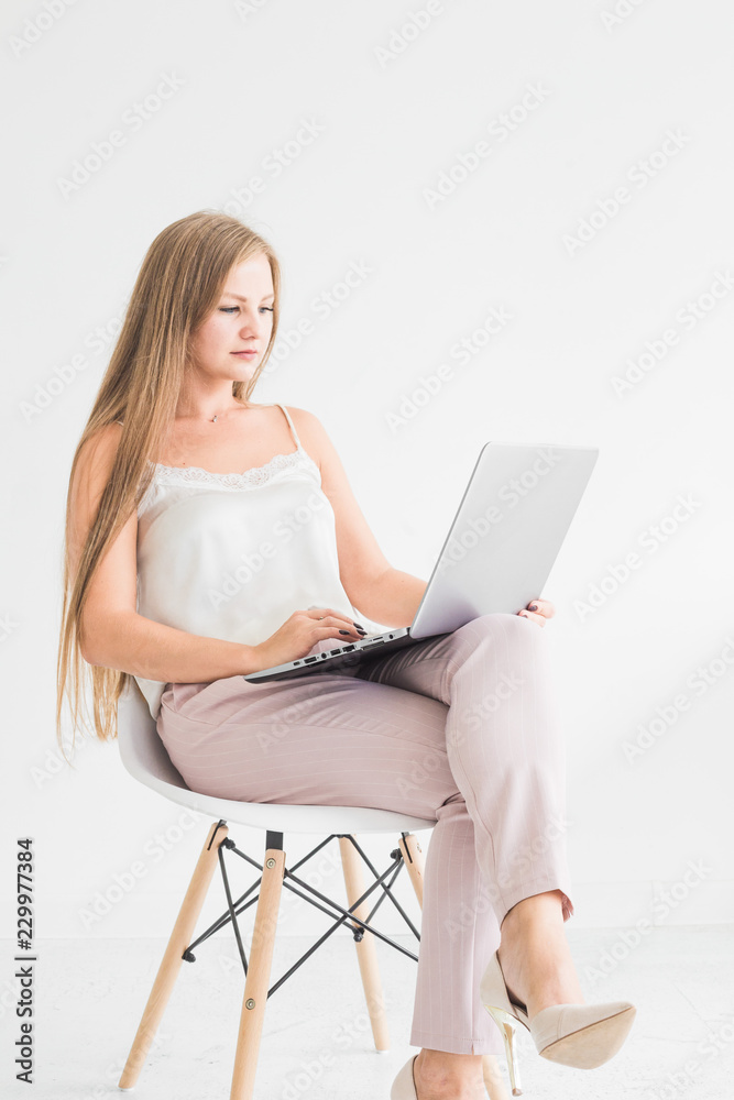 Naklejka premium Portrait of a young blonde girl sitting on a chair against a white wall and working at a laptop