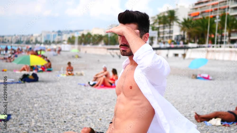 Attractive muscular young man undressing, unbuttoning and opening white shirt, revealing naked ripped torso and muscles on chest, abs, arms while looking around a beach