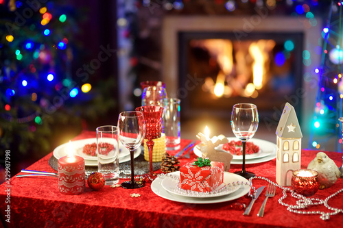 Beautiful table setting for Christmas party or New Year celebration at home. Cozy room with a fireplace and Christmas tree in a background. © MNStudio