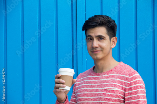 A handsome Latino man in a trendy striped sweater and with modern hair style holding a takeaway coffee cup, with a skeptical quizzical look, against a bright blue metal background. Room for text copy