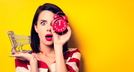 portrait of the beautiful young woman with cart for shopping and red alarm clock on the yellow background