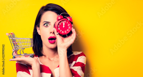 portrait of the beautiful young woman with cart for shopping and red alarm clock on the yellow background
