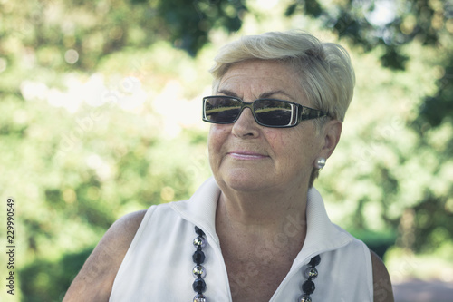Portrait of elder blonde woman with dark sunglasses on sunny day in the park. Pretty senior lady closeup posing with sleeveless white shirt