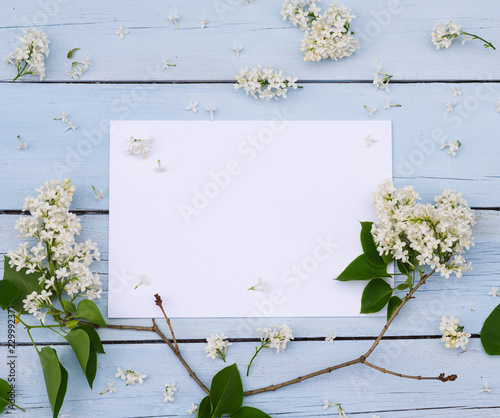 Lilac spring flowers with blank paper for greeting message on a light blue shabby wooden background. Vintage Floral mock up with white flowers. Copy space