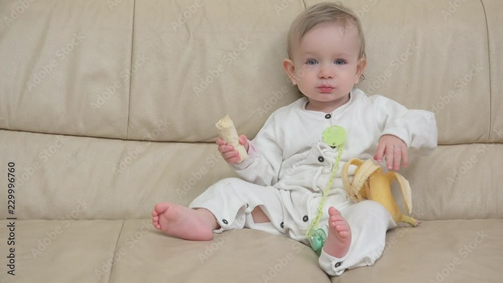 Little baby sits on sofa and eats a banana Little baby sits on sofa and eats a banana