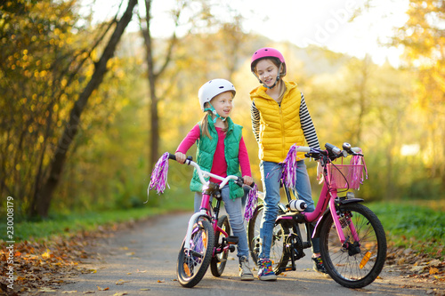 Cute sisters riding bikes in a city park on sunny autumn day. Active family leisure with kids. Children wearing safety hemet while riding a bicycle. © MNStudio