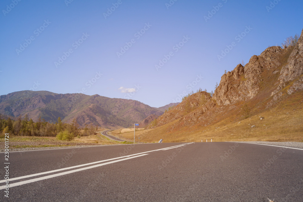 The asphalt track goes in the distance. Lower view of the road surface with white markings. Along the mountain road winds through the landscape. Landscape of Altai Mountains in Russia. The asphalt track goes in the distance. Lower view of the road surface with white markings. Along the mountain road winds through the landscape. Landscape of Altai Mountains in Russia.