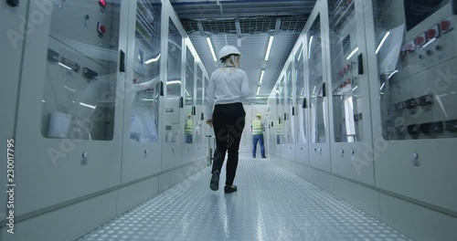 Back view of caucasian woman in hardhat walking in control center hallway of solar power station with colleagues