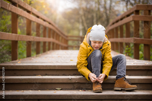 Adorable kid boy sat down to tie his shoelaces while walking in a city park on a cold autumn day. A child learns to tie shoelaces