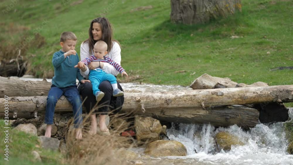 Happy young mother , baby child and big son sit on tree timber, throwing stones Happy young mother , baby child and big son sit on tree timber, throwing stones