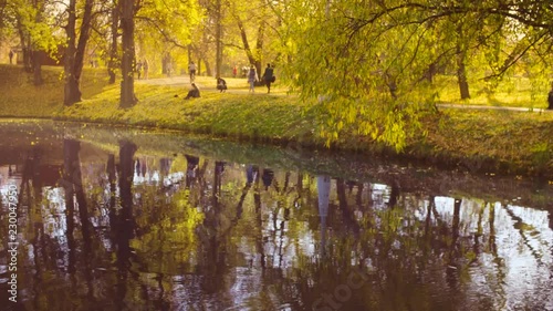 Autumn landscape, lake in the park, colorful trees reflected in the water, unrecognizable people walking on the background.