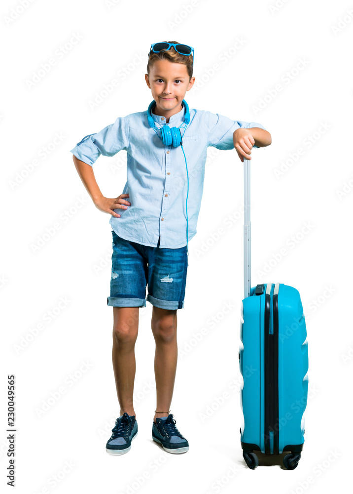 Full body of Kid with sunglasses and headphones traveling with his suitcase posing with arms at hip on isolated white background Full body of Kid with sunglasses and headphones traveling with his suitcase posing with arms at hip on isolated white background