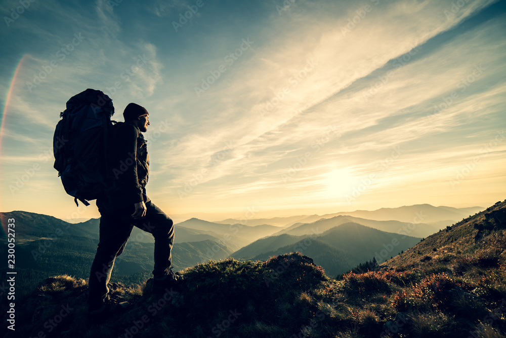 The man standing with a camping backpack on a rock with a picturesque sunset