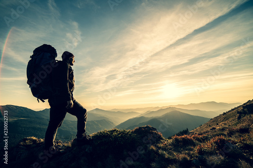 The man standing with a camping backpack on a rock with a picturesque sunset