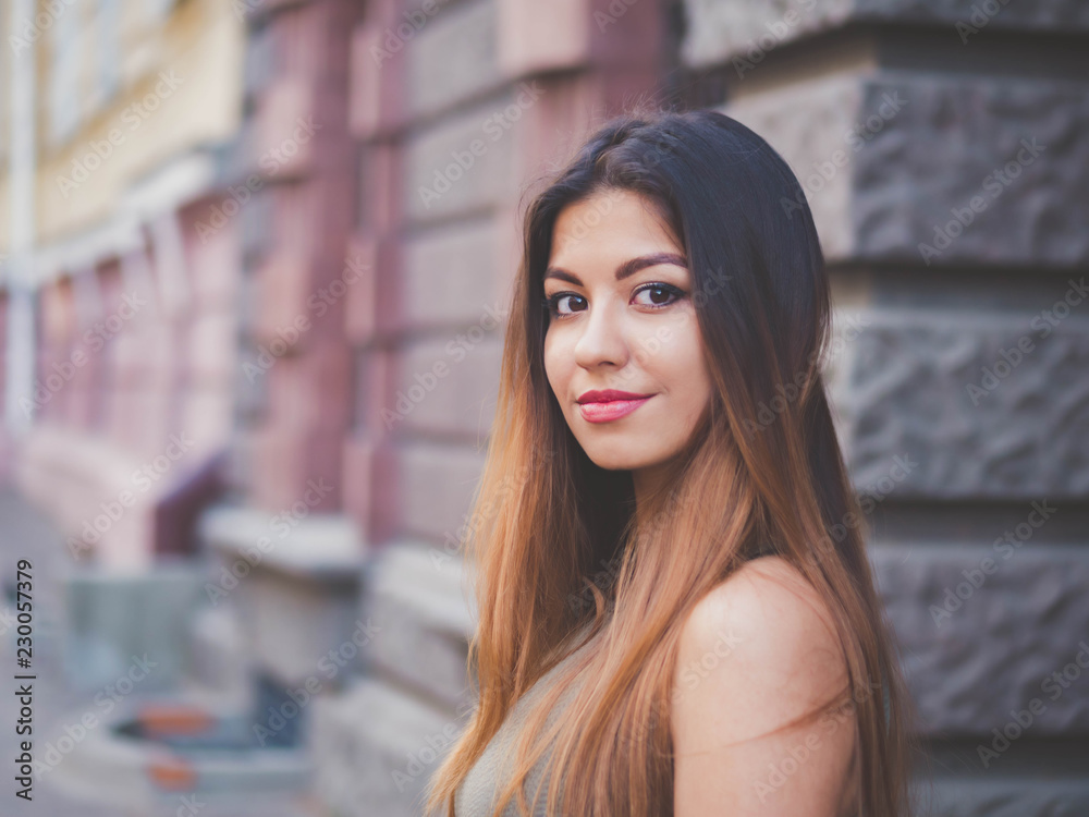 Portrait of young attractive girl looks into the camera with smile. Woman with oriental face, brown eyes and stylish ombre dyed long haistyle on architecture background. Street style