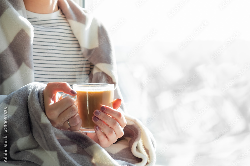 Woman's hands holding hot steaming drink. Female covered in throw blanket sits by the window with cup of hot cocoa Woman's hands holding hot steaming drink. Female covered in throw blanket sits by the window with cup of hot cocoa