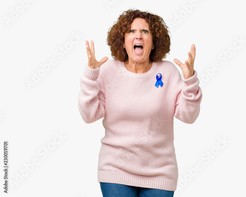 Middle ager senior woman wearing changeable blue color ribbon awareness over isolated background crazy and mad shouting and yelling with aggressive expression and arms raised. Frustration concept.