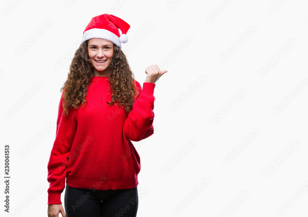 Young brunette girl wearing christmas hat over isolated background smiling with happy face looking and pointing to the side with thumb up.