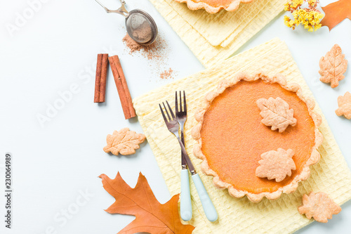 Pumpkin pie with cinnamon and cookies on yellow napkins on pastel blue background with autumn yellow leaves.