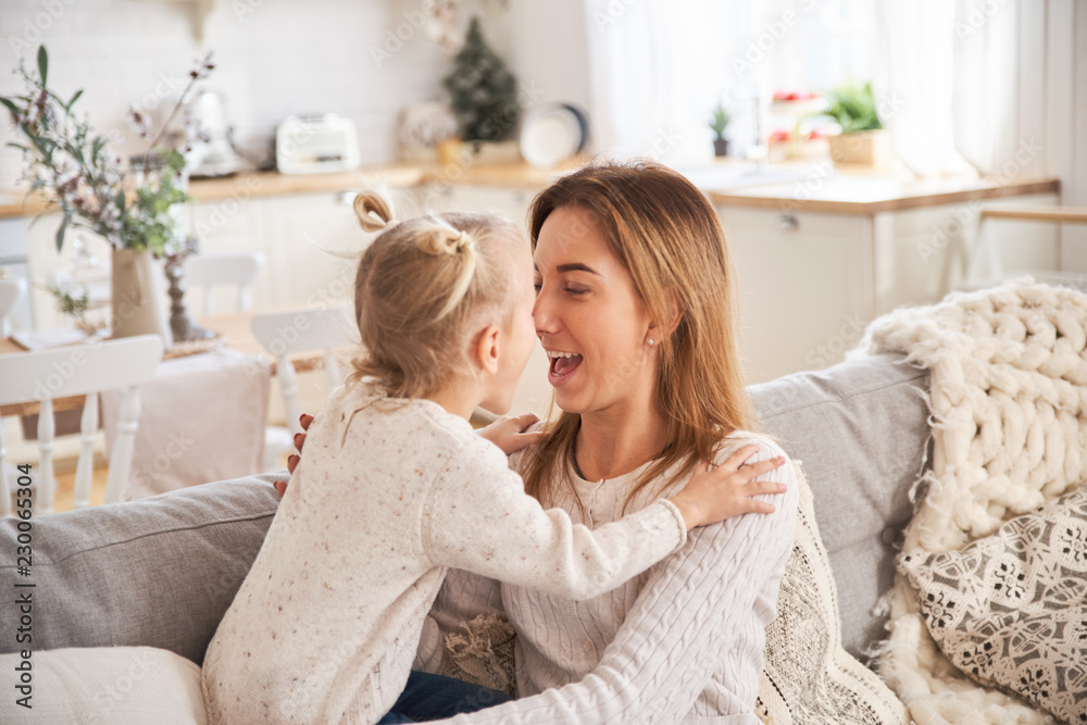 Beautiful mother woman in white sweater hugs and kisses her little daughter. Cheerful playful mood and love inside the family. Bright cozy home environment