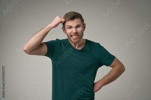 An attractive unshaven young male has desperate appearance, clenches his teeth from disappointment. An angry businessman raises his fist, knocks on head, does not control his negative emotions