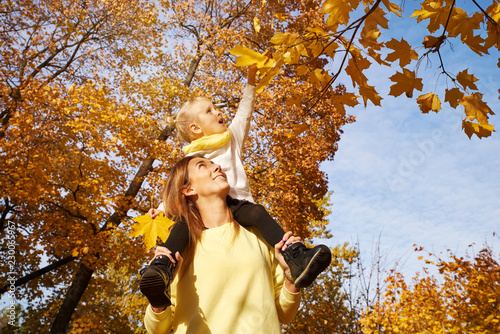 Girl sitting on the neck of the mother and stretches to autumn leaves. Beautiful autumn photo of a friendly family outdoors