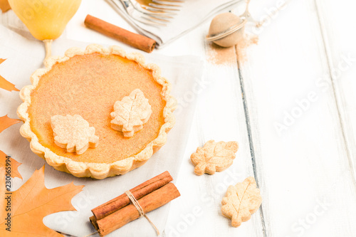 Pumpkin pie with cinnamon and cookies on gray napkins on white wooden background with autumn yellow leaves.