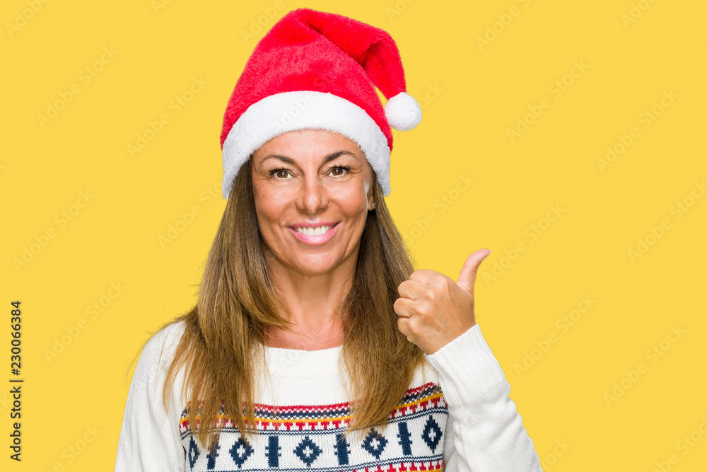Middle age adult woman wearing winter sweater and chrismat hat over isolated background doing happy thumbs up gesture with hand. Approving expression looking at the camera with showing success.